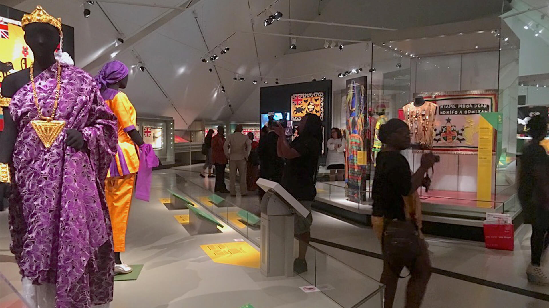 Visitors walking past several display cases containing Ghanese artifacts in an exhibition gallery space.