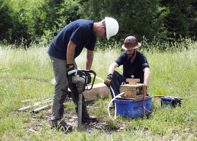 Two men with hard hats and drilling equipment in grassy field drilling into ground.