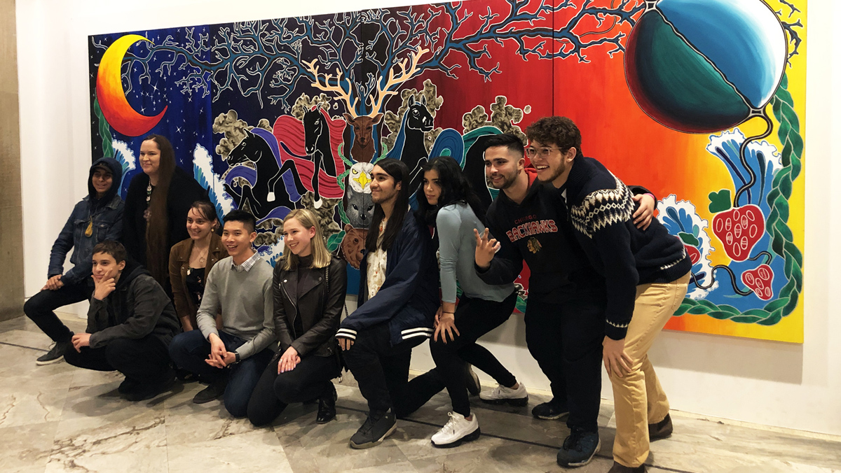 Nine Youth Cabinet members pose in front of a large mural on the wall with ROM Indigenous Knowledge Resource Teacher Leslie McCue.