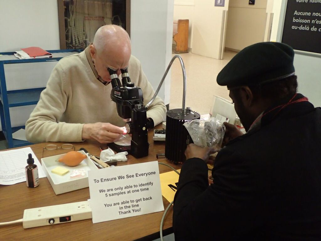 A bald man with a ring of white hair in a beige sweater studies an item under a microscope while another man in a black coat and beret is across the table, looking at another item inside a Ziploc bag