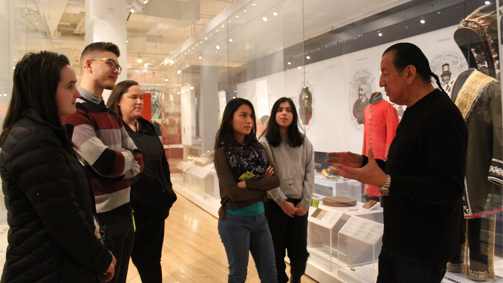 Indigenous man standing in front of a display case containing Indigenous objects with a small group of teenage students.