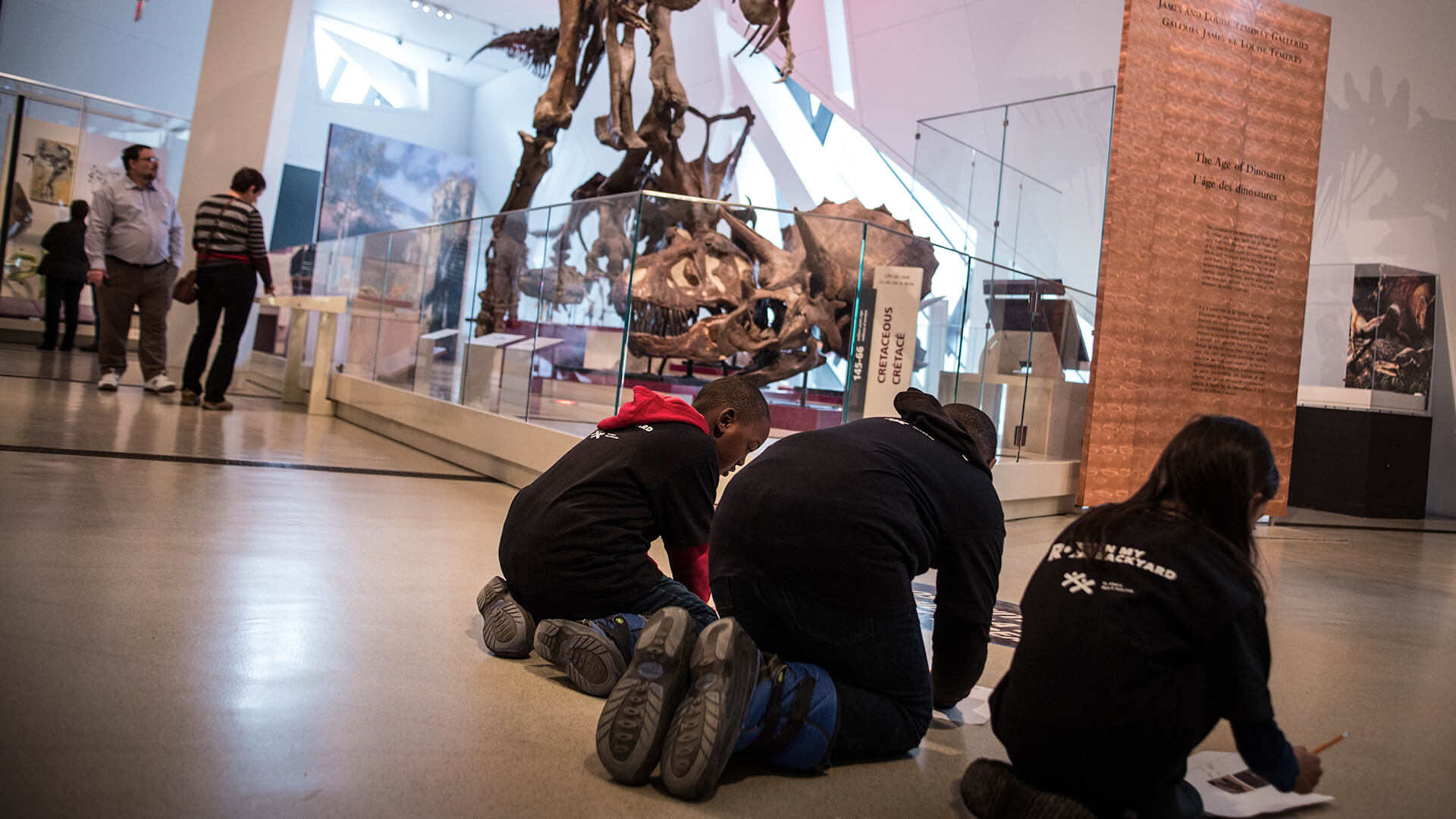 Three young students crouched on the floor in front of a dinosaur display case filling in worksheets.